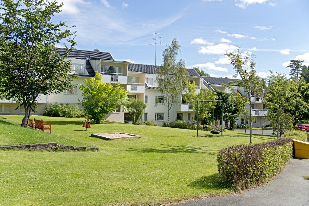 Bostadsområde med vita lägenhetshus, gräsmatta, träd och en lekplats med gungställning.